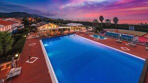 Ein Swimmingpool mit roter Liegefläche, umgeben von Gebäuden und Palmen, vor einem farbenprächtigen Sonnenuntergangshimmel.