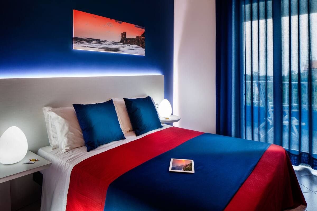 Una habitación de hotel moderna con una cama grande, una mesita de noche con lámpara, cortinas y vistas a la ciudad.