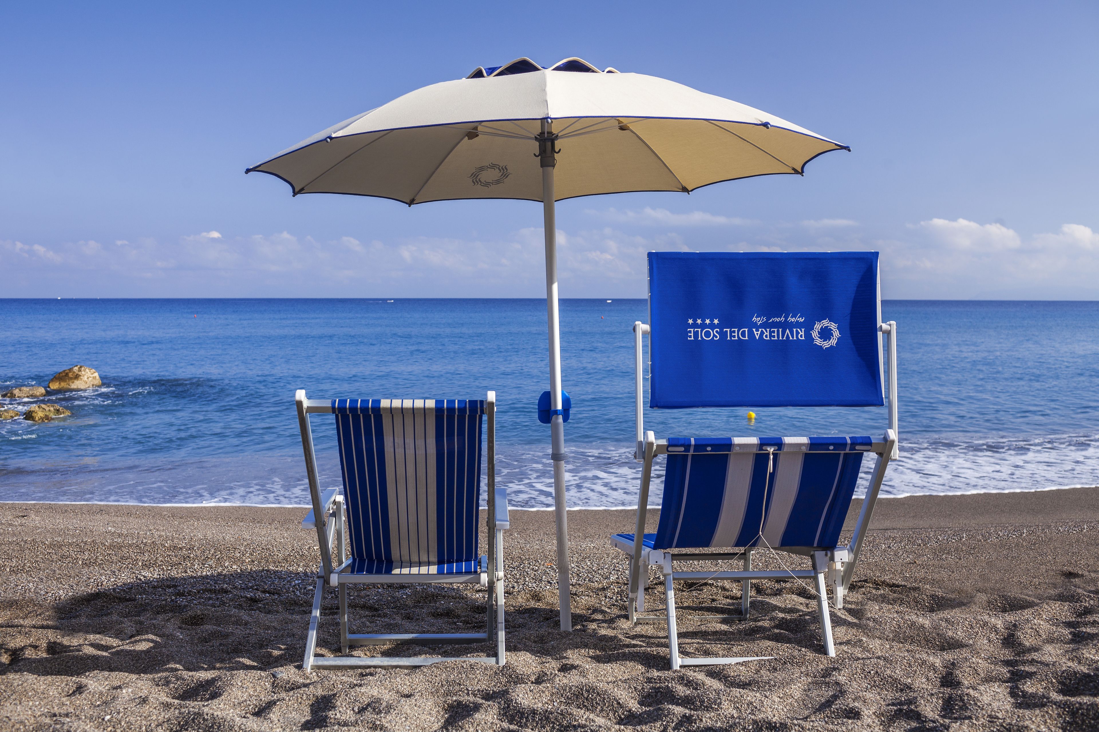 Two beach chairs with a beach umbrella and a blue towel with text, positioned on a sandy beach with the ocean in the background.
