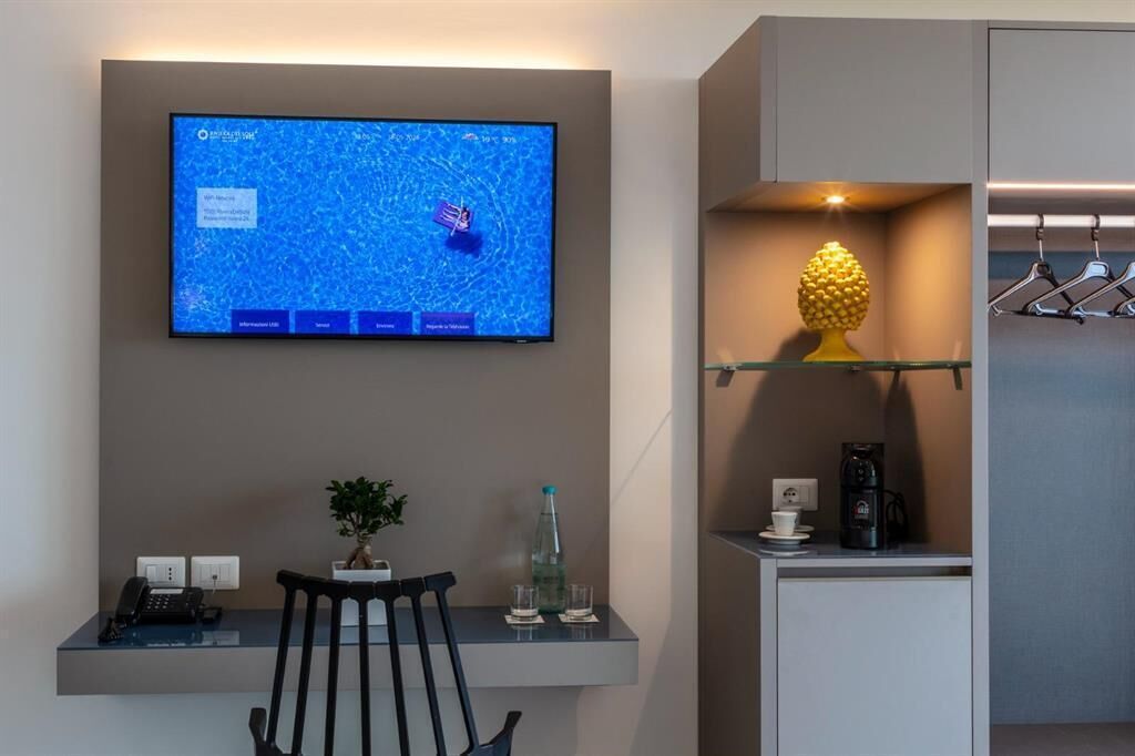 Ein modernes Hotelzimmer mit einem Wand-Fernseher, der eine Karte zeigt, einem kleinen Schreibtisch mit Telefon, einem Sessel, einer Topfpflanze, einer Kaffeemaschine und einem Regal mit einer Dekoration und Kleiderbügeln.
