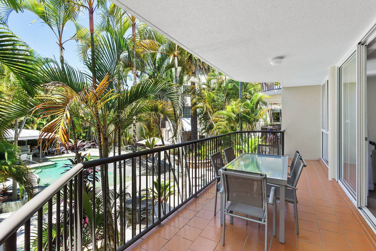 Ein Balkon mit Glastisch und Stühlen, umgeben von Palmen und einem Pool.