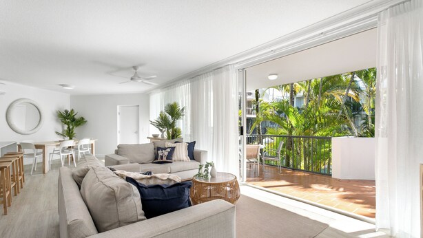 Een ruime woonkamer met een grote bank, een eethoek en een balkon met uitzicht op palmbomen.