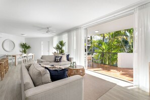 A spacious living room with a large sofa, a dining area, and a balcony with a view of palm trees.