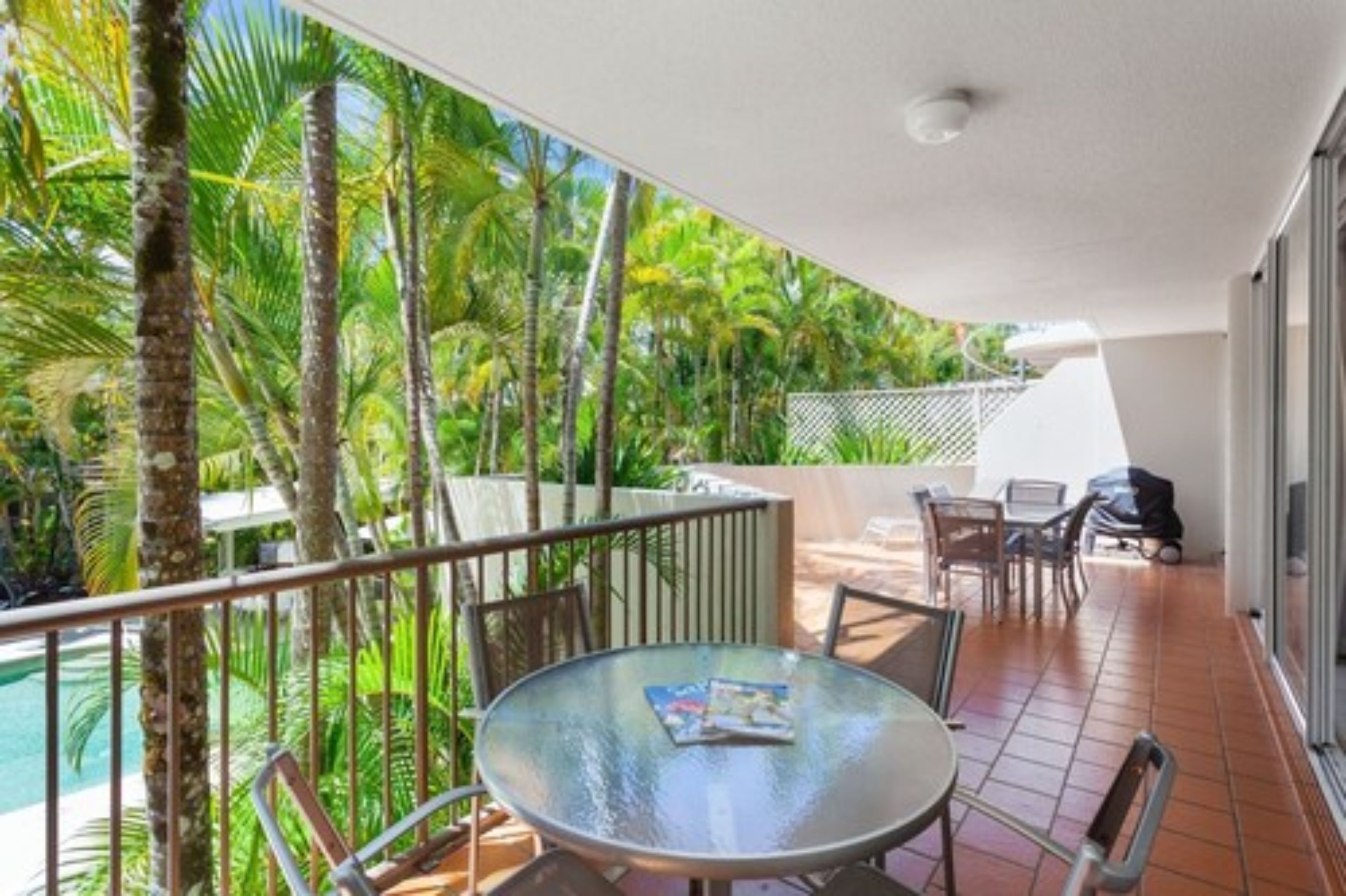 A balcony with a glass table, chairs, and a pool view.