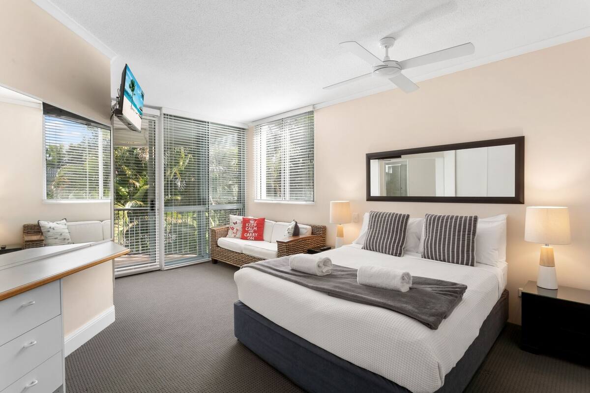 Ein modernes Hotelzimmer mit einem großen Bett, einem Deckenventilator, einem Fernseher und einem Balkon mit großen Fenstern.