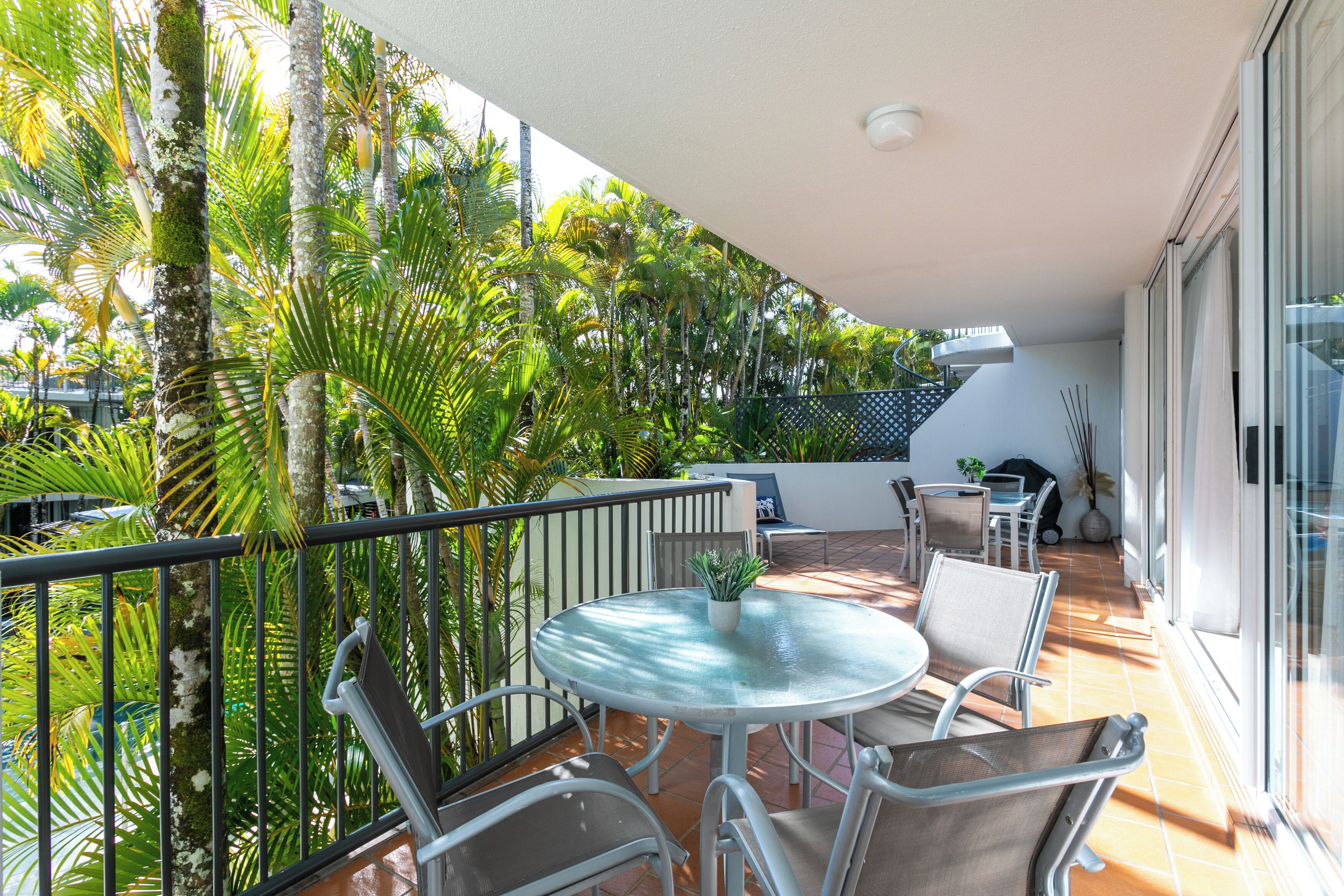 Un balcon doté d’une table ronde en verre et de chaises en métal, entouré d’un luxuriant écrin de palmiers.