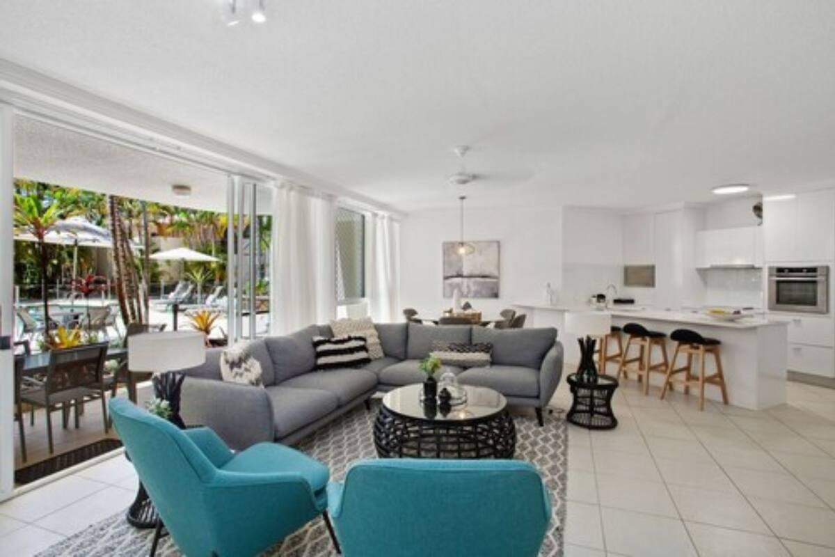 Ein modernes Wohnzimmer mit einer grauen Couch, türkisfarbenen Sessel und einem runden Couchtisch, sowie einer Küchenzeile mit weißen Schränken und einer Bar.
