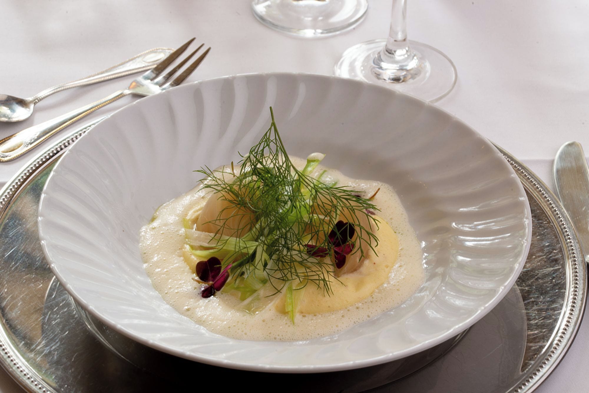 Un plato blanco con un guiso cremoso, decorado con eneldo y flores comestibles, sobre una bandeja de plata.