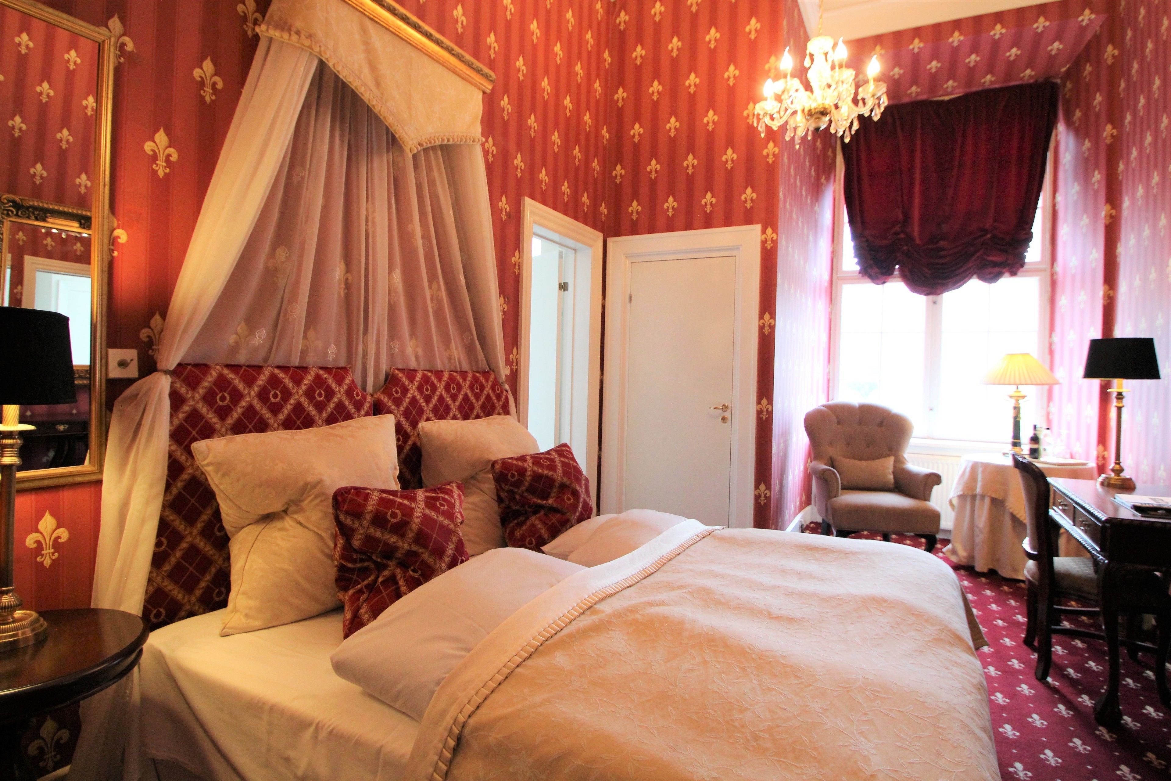 A bedroom with a four-poster bed, a chandelier, a desk with a lamp, and a chair.