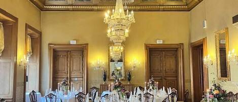A formal dining room with ornate ceiling and chandeliers, wooden chairs, and tables set for a meal.