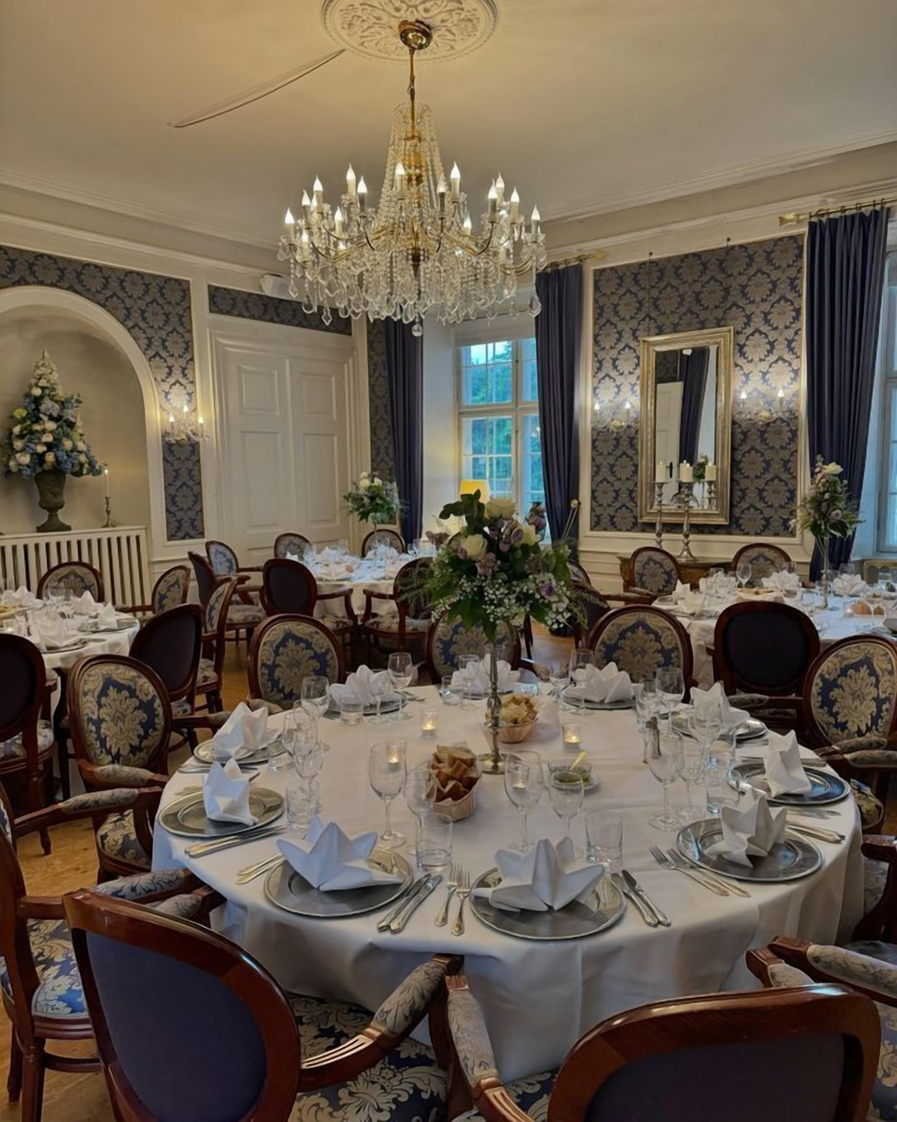 A formal dining room with round tables set for a meal, elegant chairs, a chandelier, and patterned wallpaper.