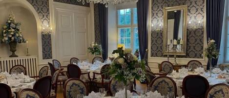 Une salle à manger formelle avec des tables rondes dressées pour le repas, des chaises élégantes, un lustre et un papier peint à motifs.