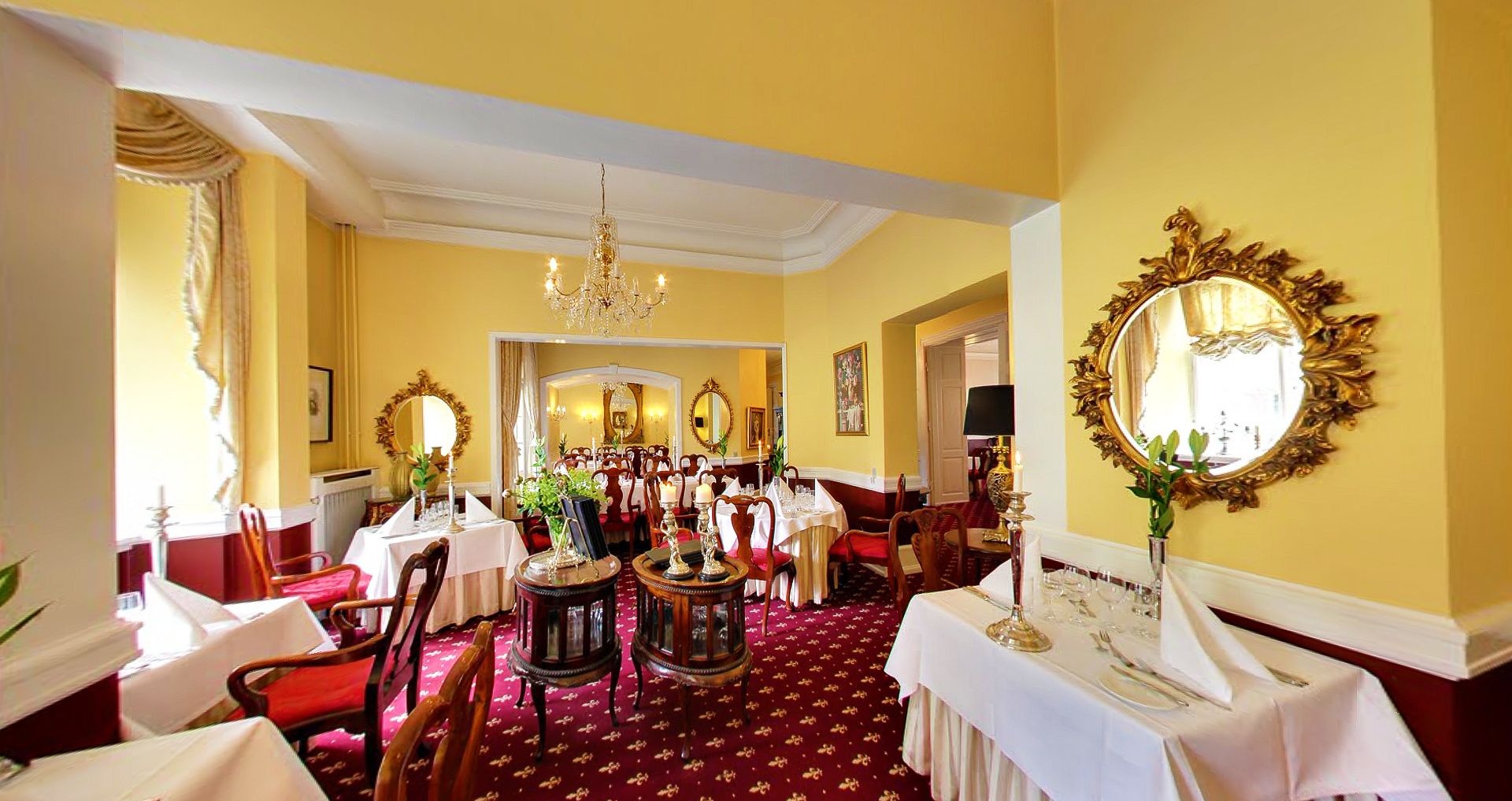 A formal dining room with multiple tables set for service, a chandelier, and ornate wall mirrors.