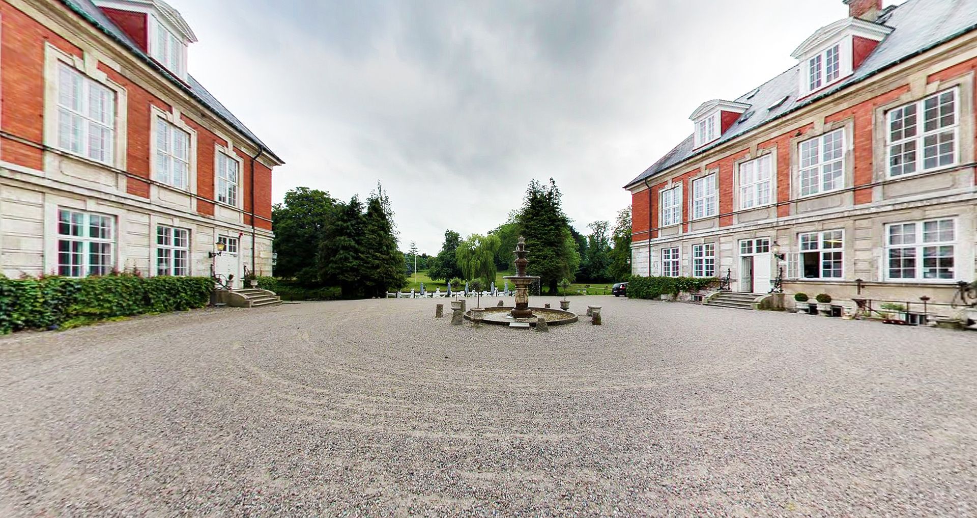 Ein Innenhof mit einem zentralen Brunnen, umgeben von zwei symmetrischen Gebäuden mit großen Fenstern und Kiesböden.