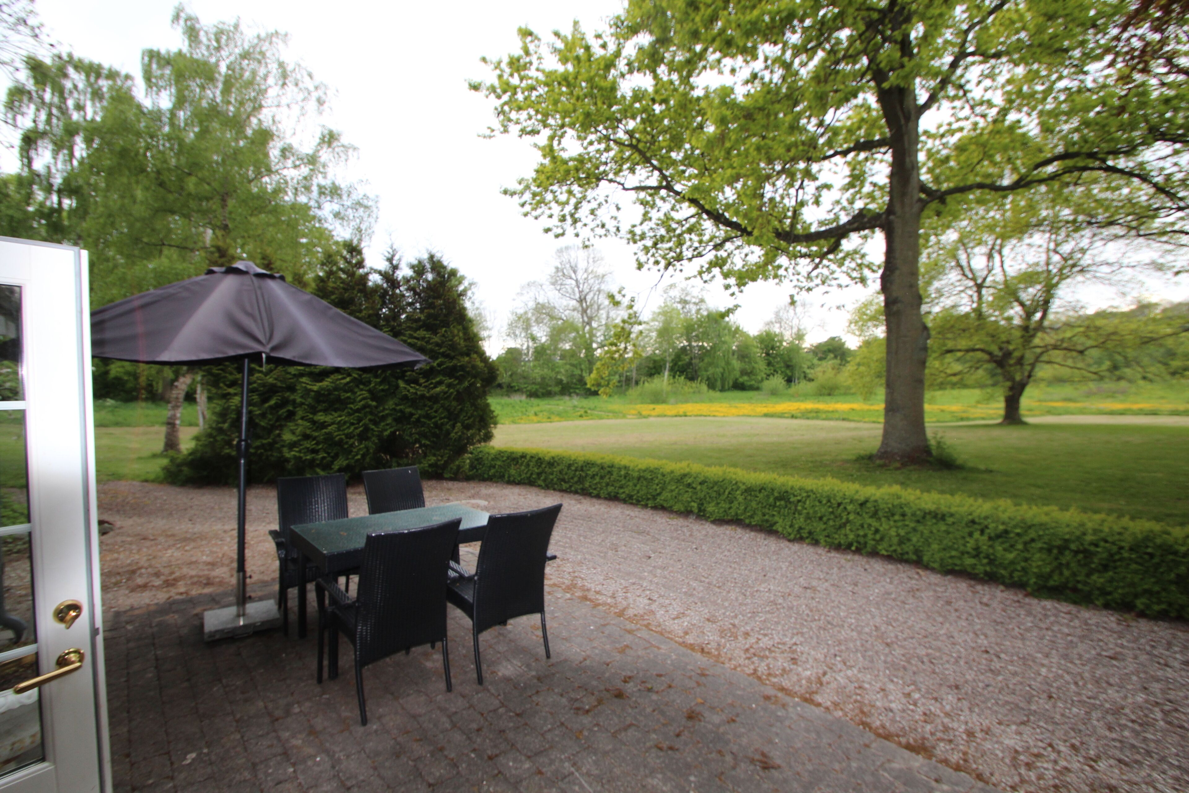 An outdoor patio with a table and chairs, a dark umbrella, and a view of a manicured lawn and trees.