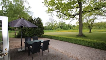 An outdoor patio with a table and chairs, a dark umbrella, and a view of a manicured lawn and trees.