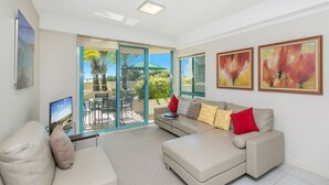 A modern living room with a beige sofa, a coffee table, and a TV. There are sliding glass doors leading to an outdoor patio with a table and chairs. - Argyle on the Park (Maroochydore)