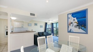 A modern kitchen with white cabinets, a dining area with a glass table and white chairs, and a large window with a view of the sea. - Argyle on the Park (Maroochydore)