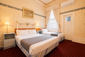 A hotel room with two beds, a nightstand, a lamp, a window with curtains, a door, and a wall-mounted picture. - Georgian Court Inn (East Melbourne)
