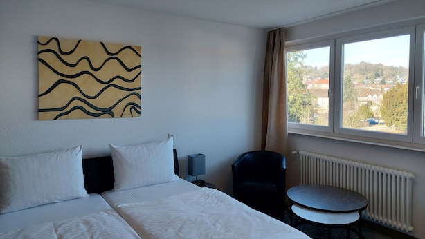 Hotelkamer met een bed, een klein tafeltje, een stoel, een radiator en een raam met uitzicht op gebouwen.