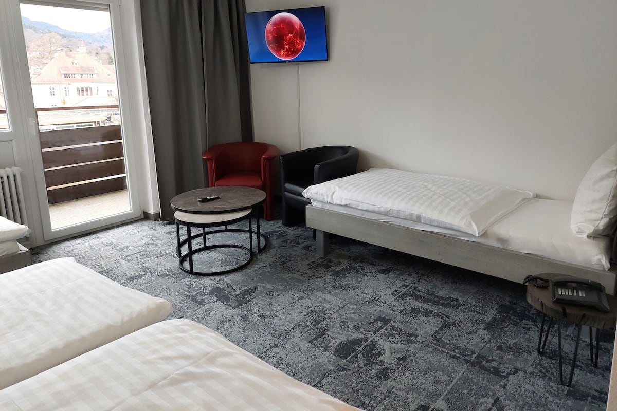 A hotel room with a bed, a small table, a red chair, a black chair, and a TV displaying a red object.