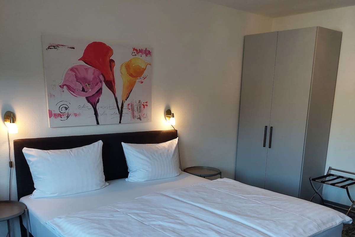 A neatly made bed with white linens, a headboard with two wall-mounted lights, a large grey wardrobe, and a wall-mounted artwork of flowers.