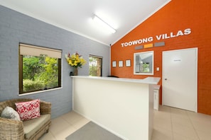 A reception area with a white counter, a wicker sofa with red cushions, a window with a view of greenery, and a wall with the text "TOOWONG VILLAS". - Toowong Villas (Toowong)