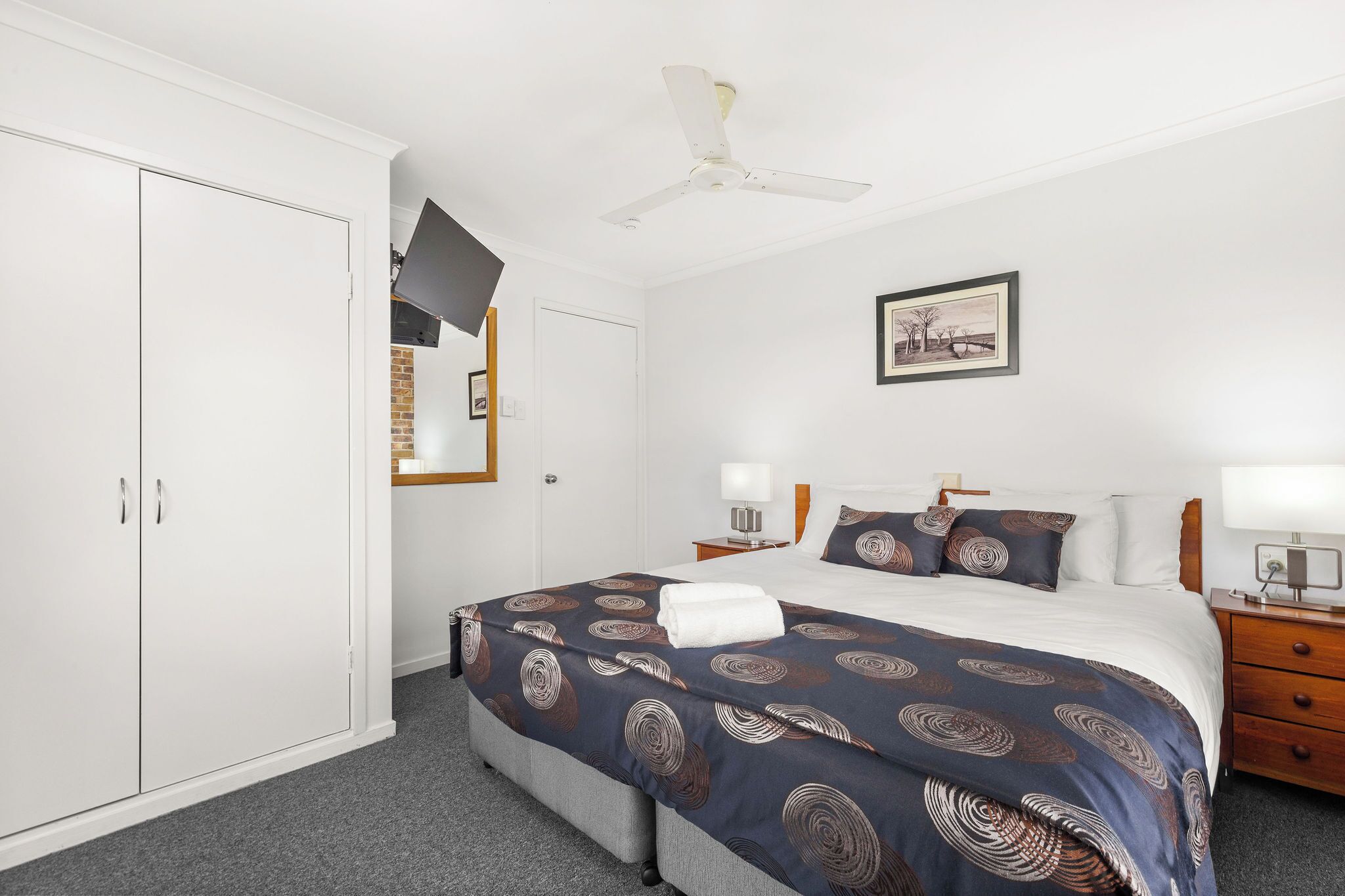 A hotel room with a bed, bedside tables, a wardrobe, a ceiling fan, and a framed picture on the wall.