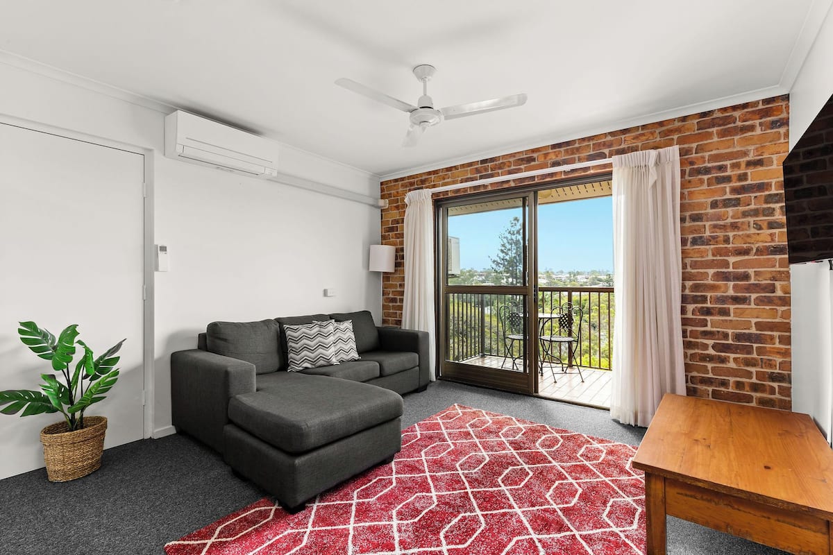 A modern living room with a grey sofa, a wooden table, a red patterned rug, and a balcony with a view.
