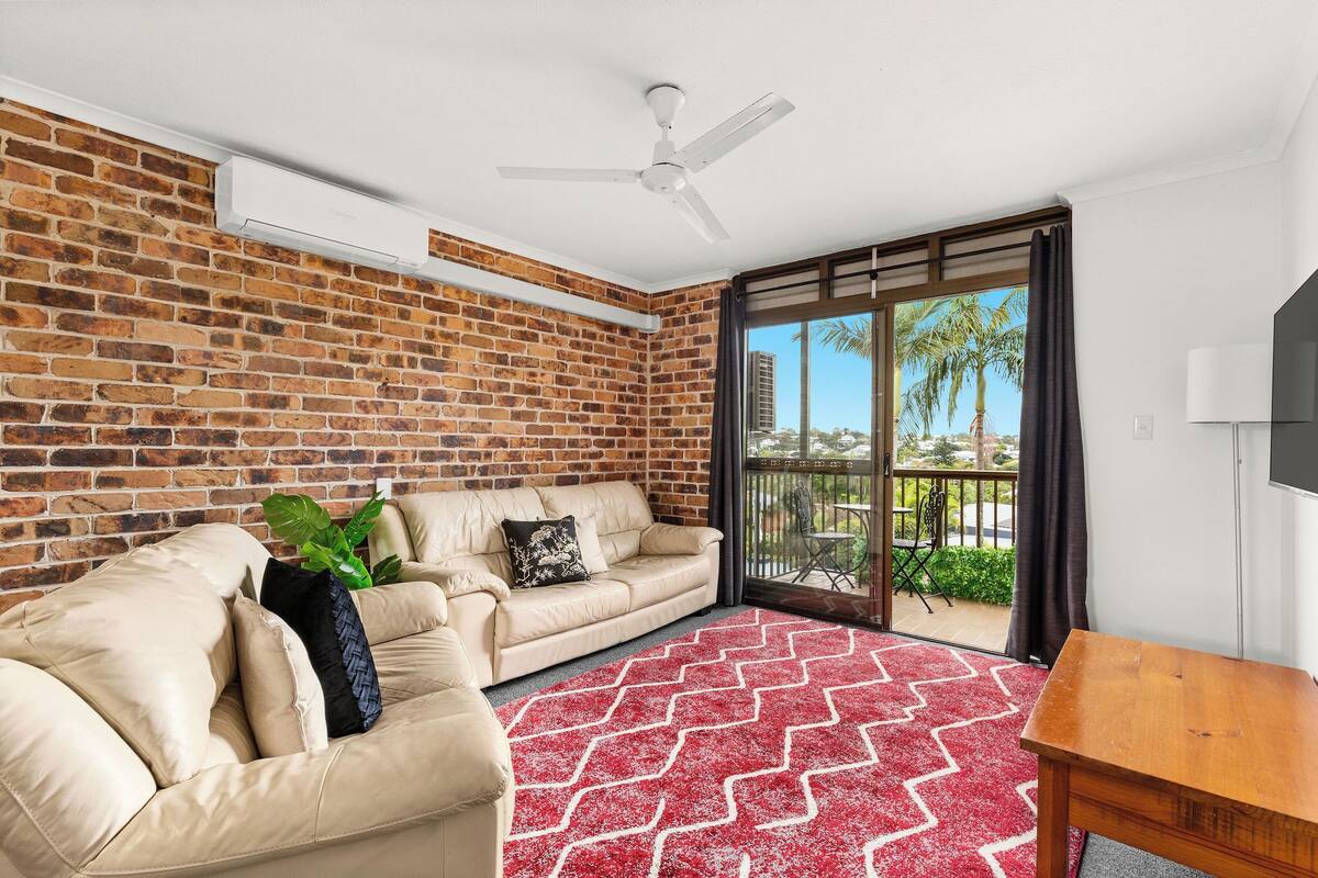 A modern living room with a brick wall, a ceiling fan, a beige sofa, a red patterned rug, and a wooden table.