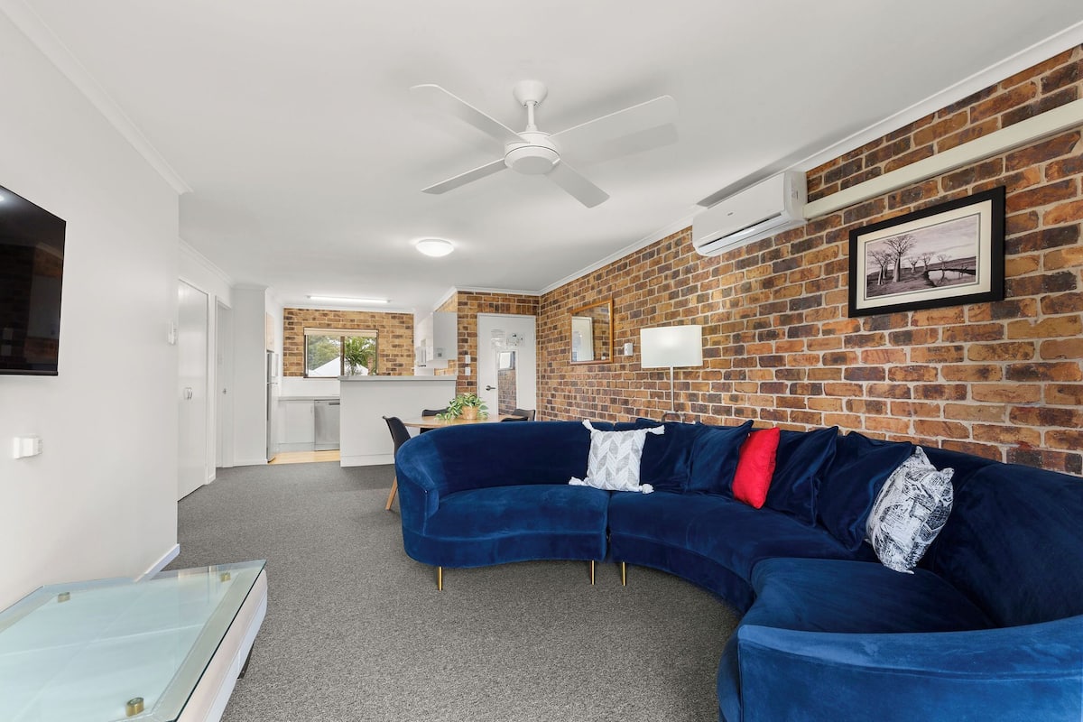 A modern living room with a curved blue sofa, a glass coffee table, and a brick wall.