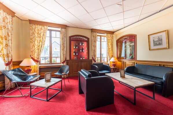 A hotel lobby with a red carpet, black leather sofas, a wooden coffee table, and a red armchair. There are two lamps on the side, and a wall with framed pictures.