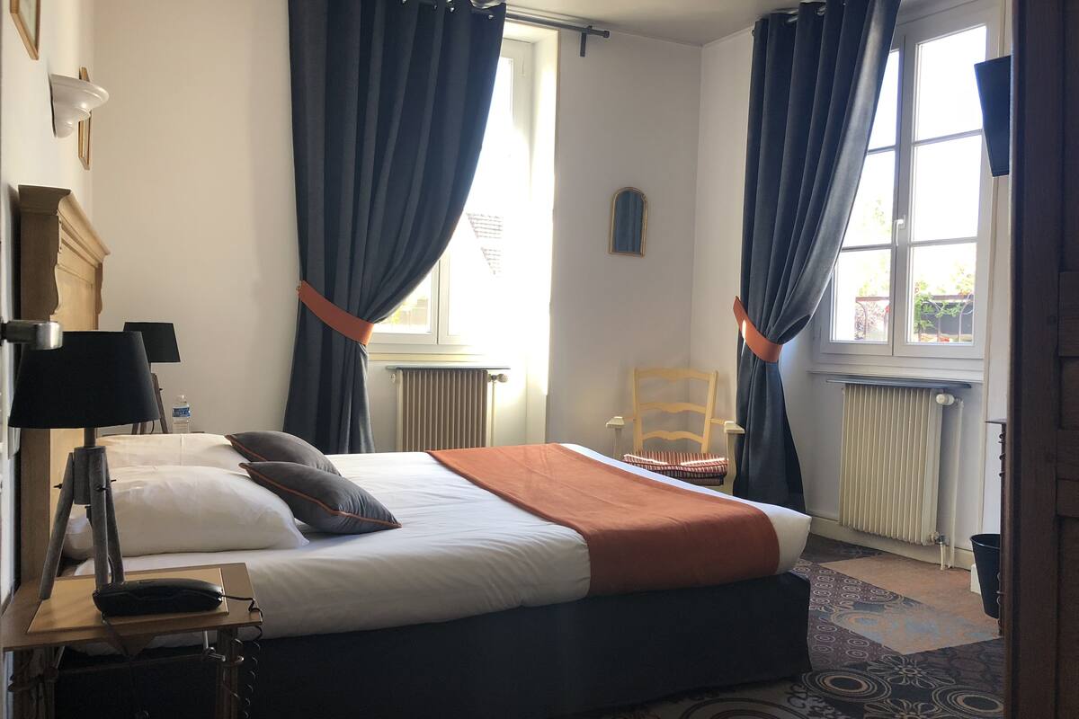 A hotel room with a bed, a desk with a lamp, a chair, and a window with curtains.