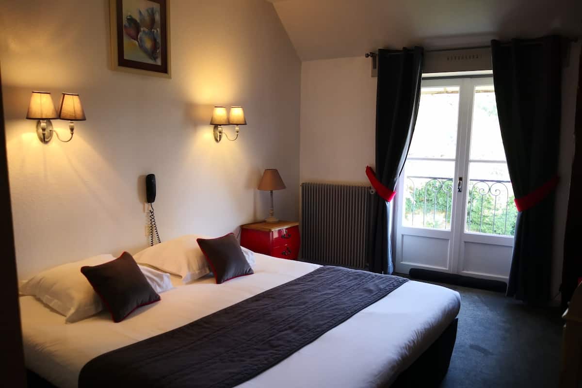 A hotel room with a large bed, two bedside lamps, a red nightstand, a telephone, a radiator, and a window with curtains.