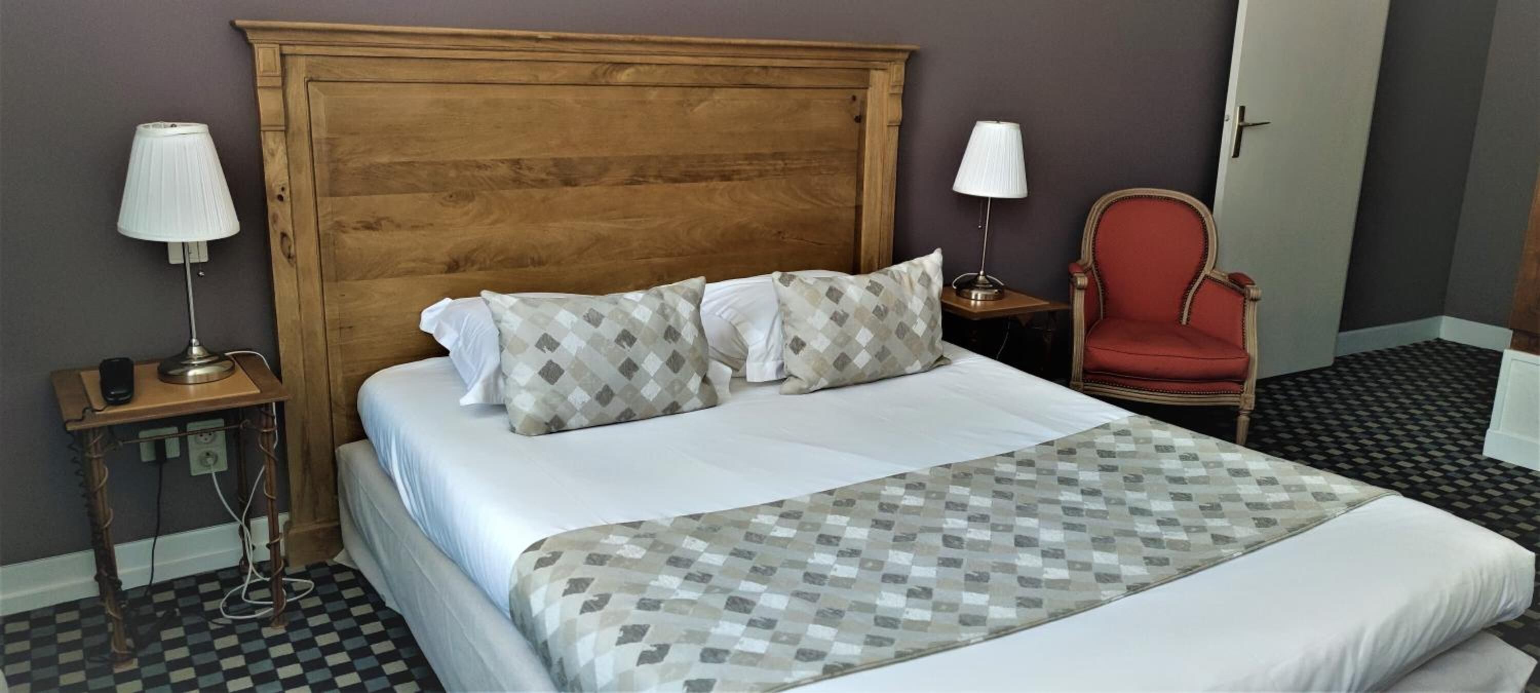 A hotel room with a large wooden headboard, two bedside tables with lamps, a red armchair, and a checkered floor.