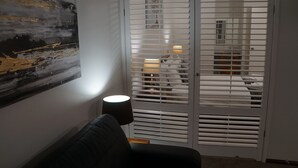 Een kamer met een zwarte bank, een witte deur met horizontale zonwering en een schilderij aan de muur.