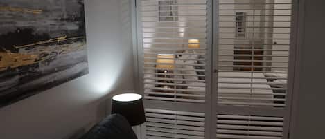 Una habitación con un sofá negro, una puerta blanca con persianas horizontales y un cuadro en la pared.