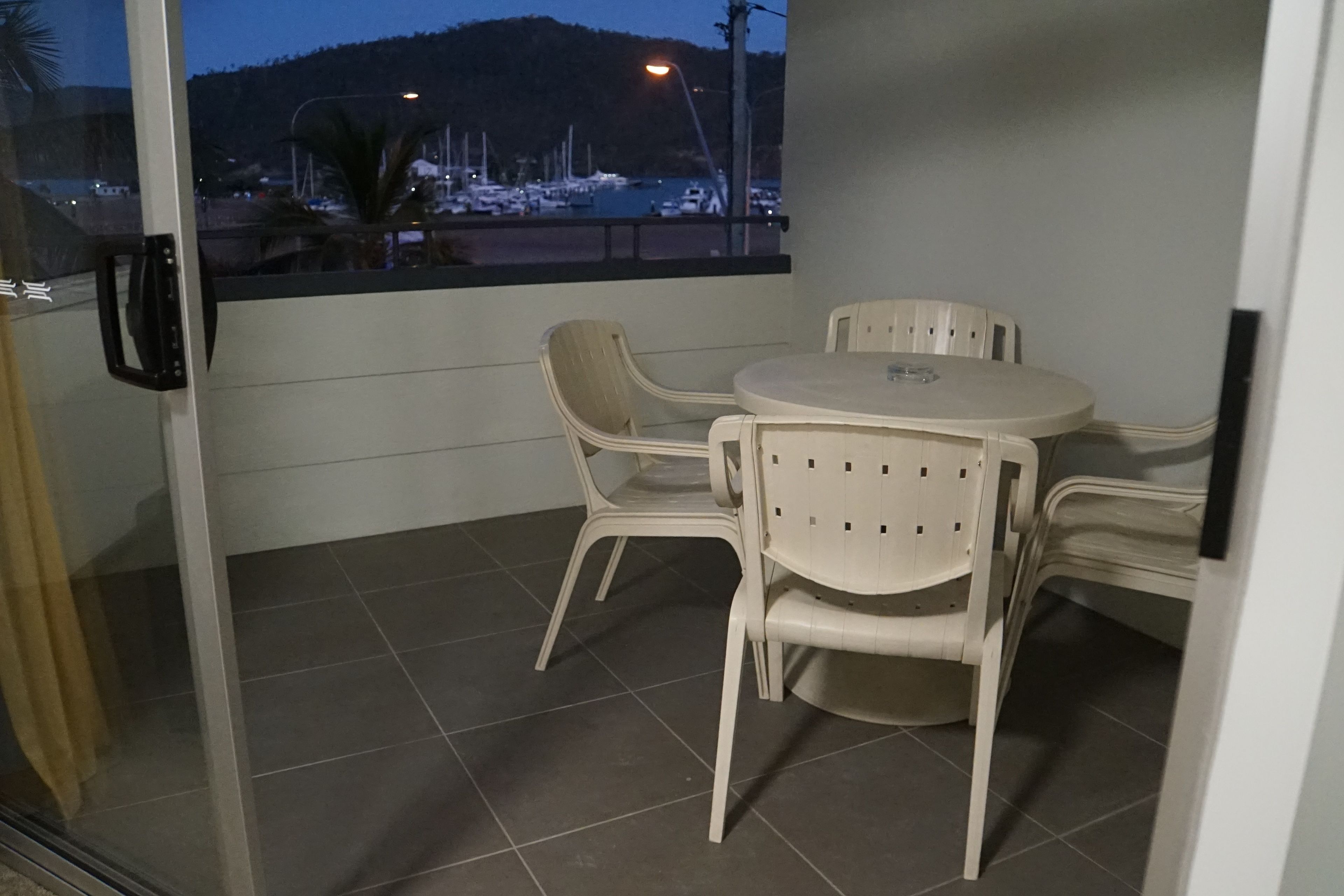 Een balkon met een glazen deur, een ronde tafel met stoelen, en een uitzicht op boten en een heuvel op de achtergrond.