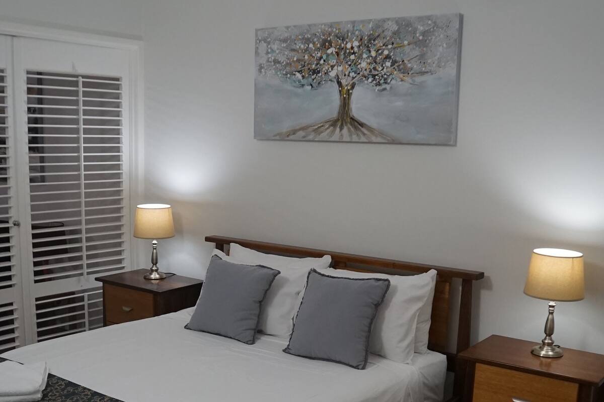Un dormitorio ordenado con una cama, dos mesitas de noche con lámparas, un ventilador de techo y un cuadro de un árbol en la pared.