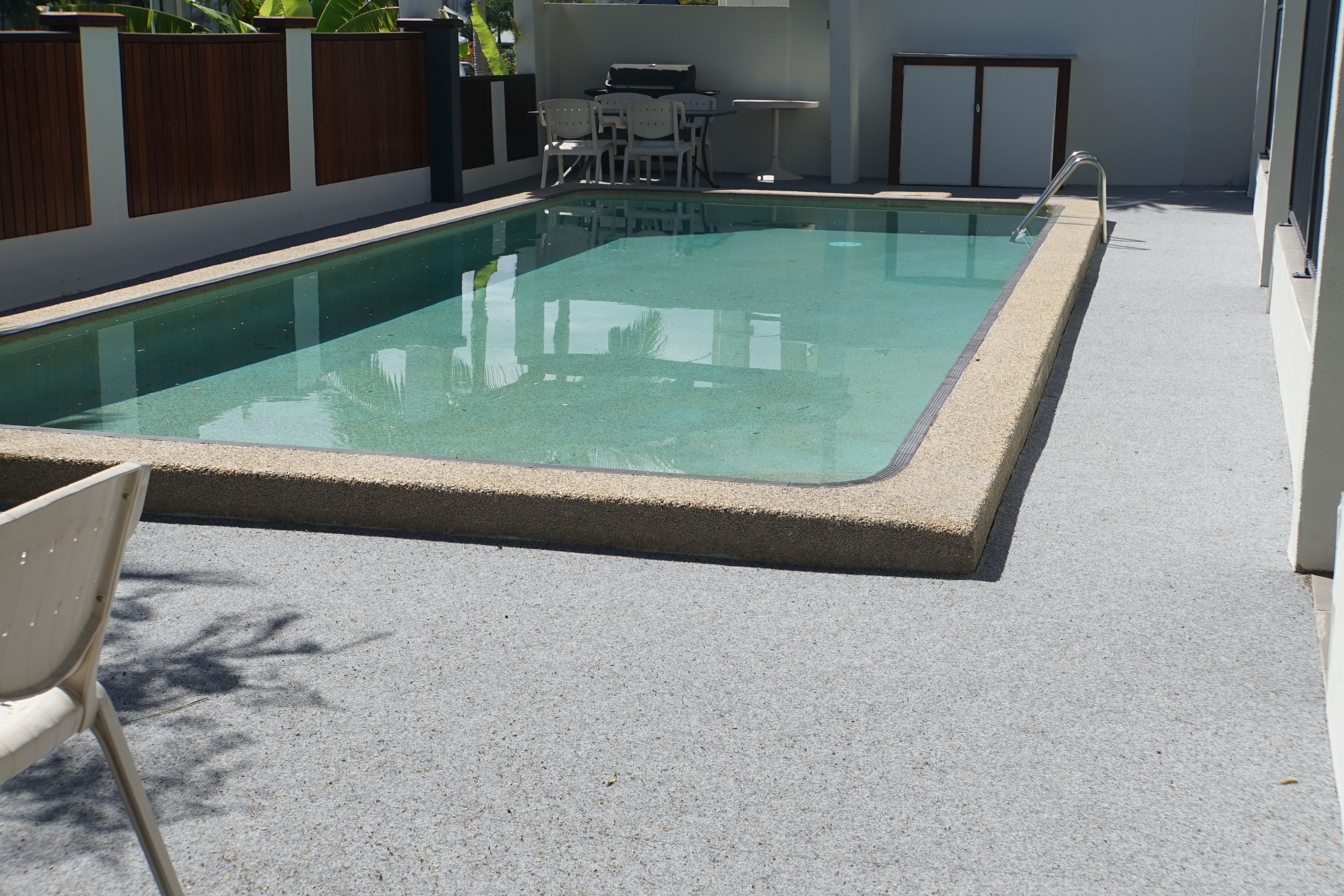 Une piscine rectangulaire bordée de béton, entourée d’une terrasse également en béton. On aperçoit une table et des chaises d’extérieur blanches, situées à gauche de la piscine.