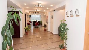 A hotel hallway with a dining area in the background, potted plants, and clocks on the wall. - Plus Hotel (Malu Mare)