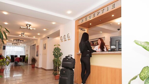 A hotel reception area with a receptionist, a desk, and a 'PLUS' sign. There are potted plants and a dining area in the background. - Plus Hotel (Malu Mare)
