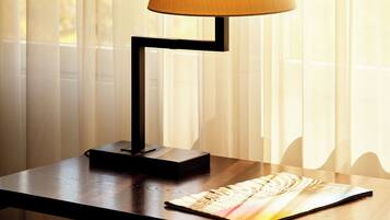 A table with a lamp, a magazine, and a chair in a room with sheer curtains.