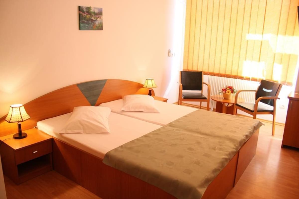 A hotel room with a bed, two bedside lamps, a chair, a small table with a vase of flowers, and a painting on the wall.