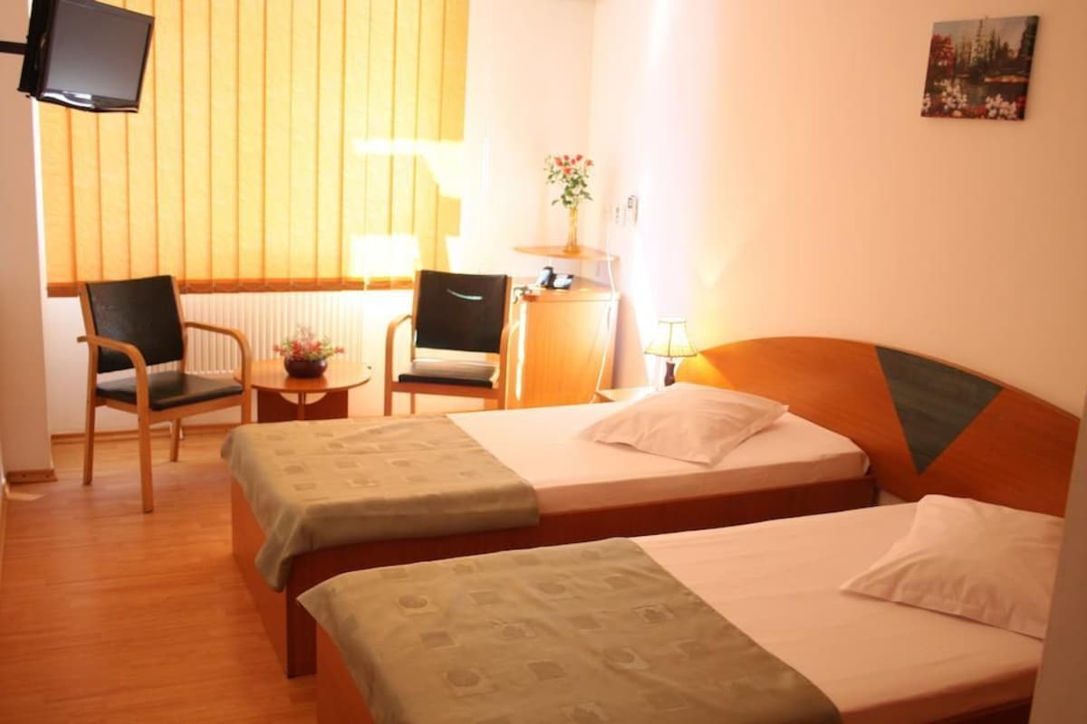 A hotel room with two beds, a desk, chairs, a TV, and a window with blinds.