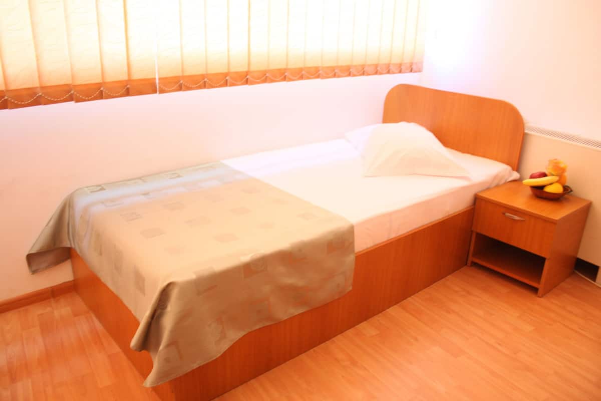 A single bed with a wooden headboard, a bedside table with a fruit bowl, and a window with blinds.