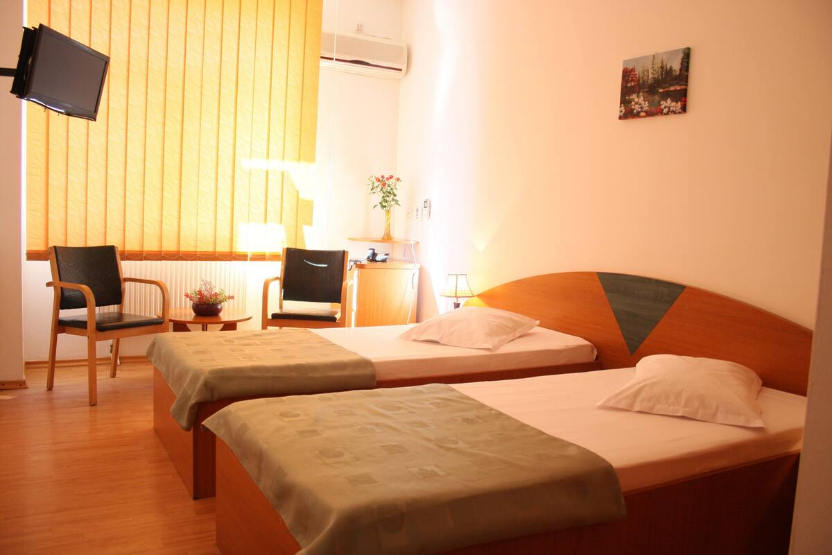 A hotel room with two single beds, a desk with a chair, a TV mounted on the wall, and a window with blinds.