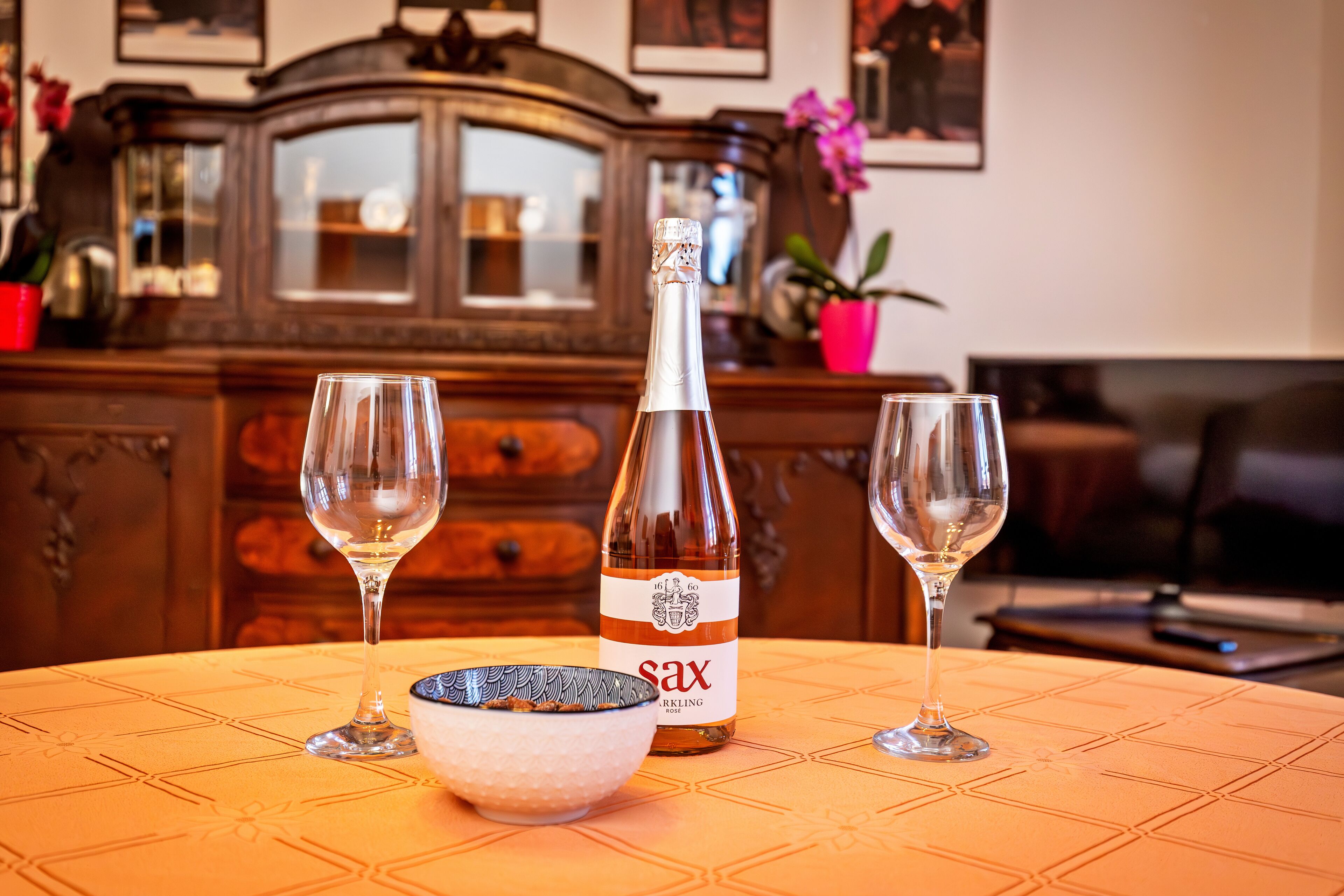 Une bouteille de vin mousseux Sax, deux verres à vin vides et un bol décoratif sur une table recouverte d’une nappe à motifs.
