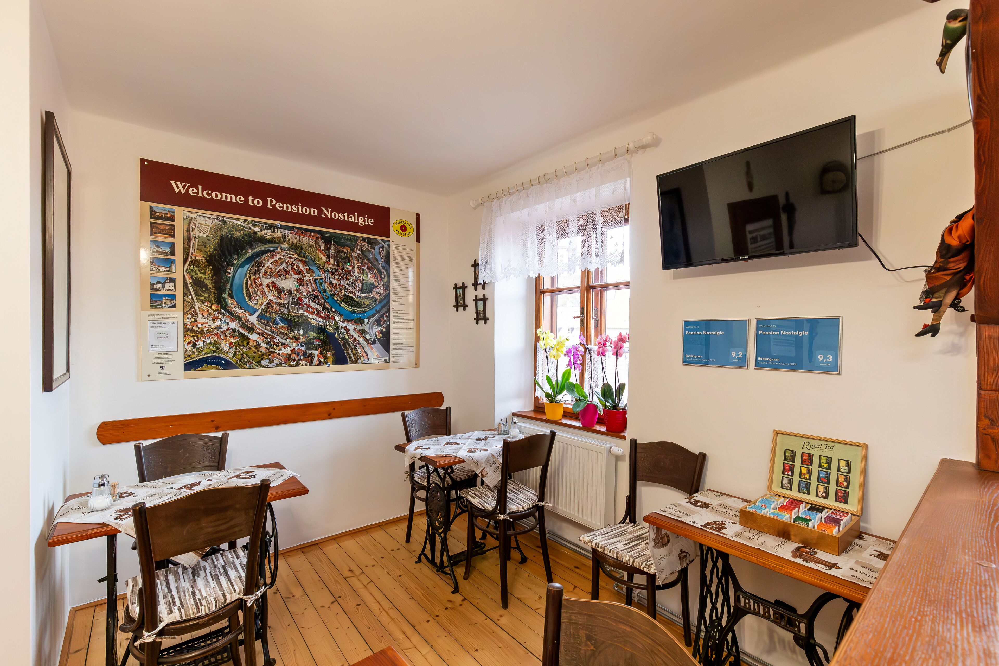 A cozy room with a wooden table and chairs, a large welcome poster, a window with flowers, and a small table with a tea display.