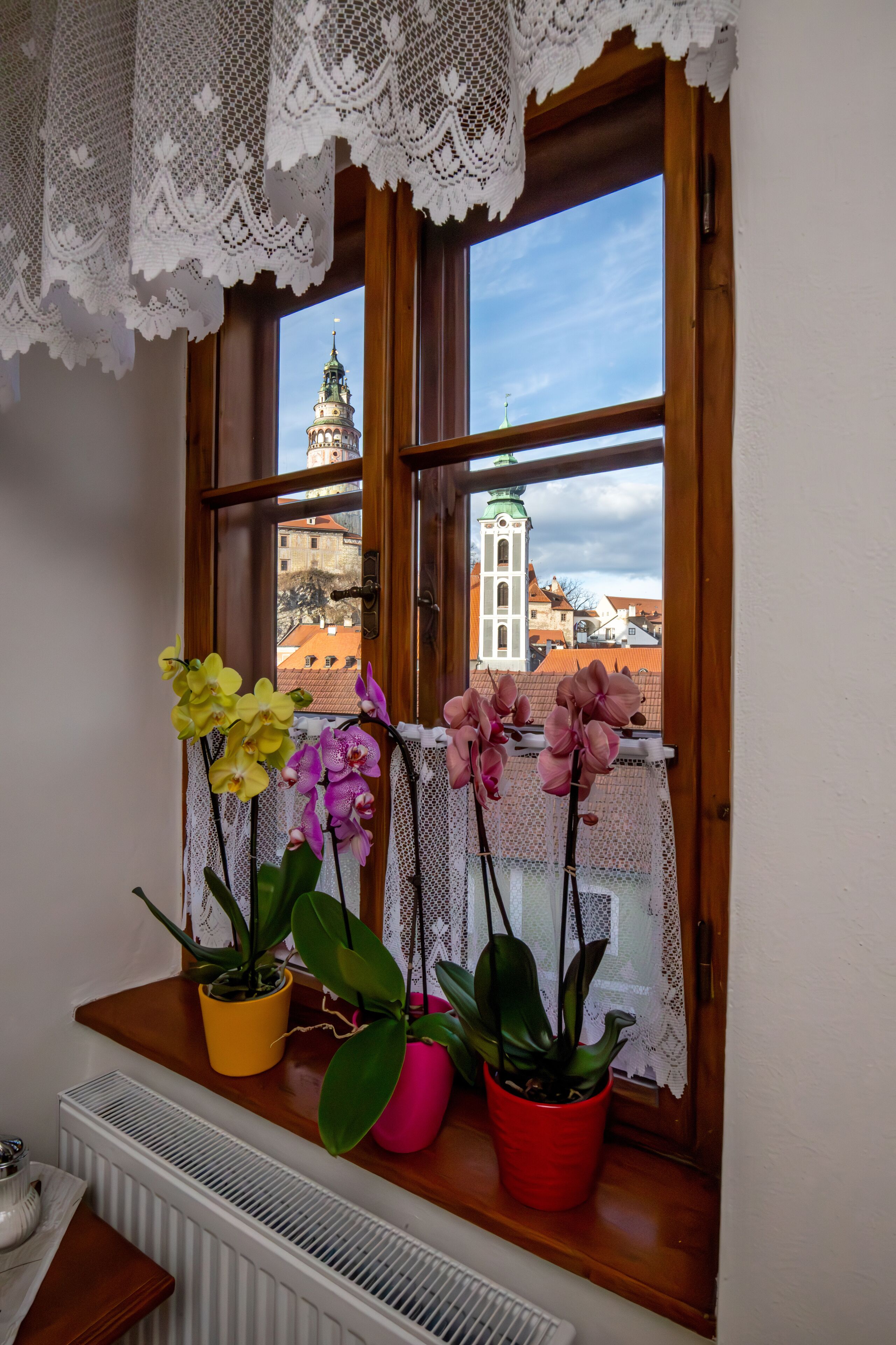 Una ventana con cortinas de encaje, tres orquídeas en macetas y vista a un edificio con una torre.